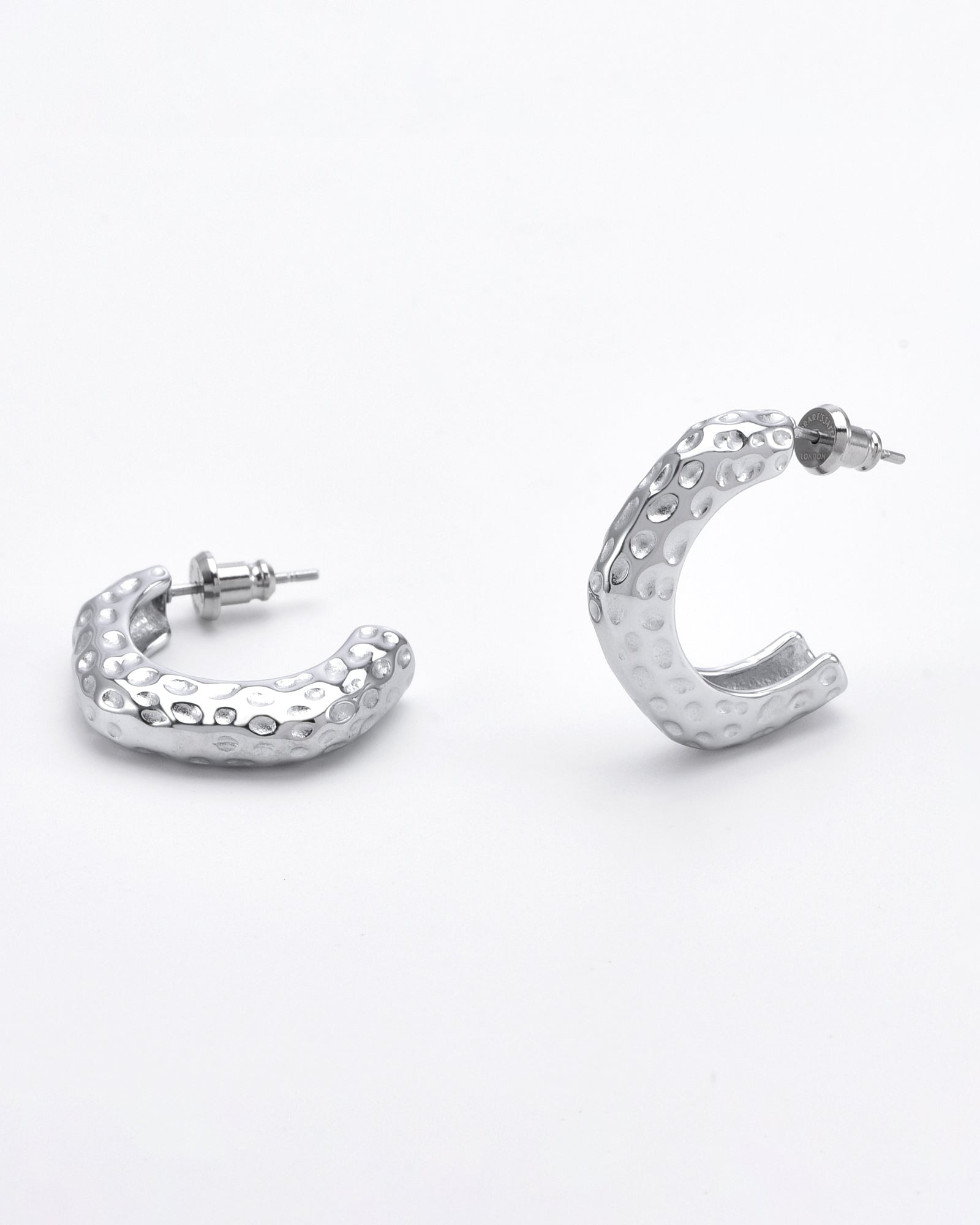 A pair of chunky, textured Mia Earrings Silver by For Art's Sake® is displayed. One earring lies flat, showing its palladium-plated surface, while the other stands upright, partially open to reveal the post and back. The background is plain and white, highlighting the earrings' intricate details.