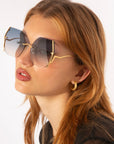 A young woman with long, light brown hair is wearing oversized, angular sunglasses with gold frames and gradient lenses. She has For Art's Sake® Mia Earrings Gold, hoop style, and is dressed in a black top. Her lips are slightly parted as she tilts her head to the side.