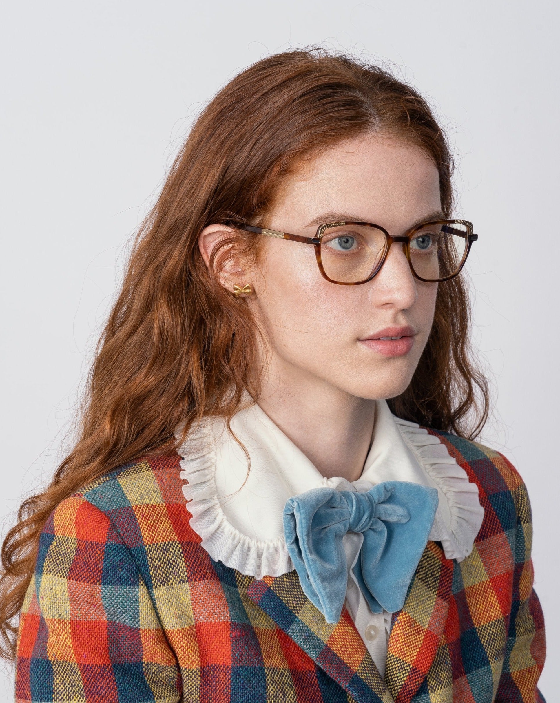 A woman with long red hair and glasses featuring Mimosa by For Art&#39;s Sake® lenses is wearing a colorful plaid jacket with a white ruffled collar shirt and a large light blue bow tie. She is looking slightly to the left against a plain white background.