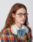 A woman with long red hair and glasses featuring Mimosa by For Art's Sake® lenses is wearing a colorful plaid jacket with a white ruffled collar shirt and a large light blue bow tie. She is looking slightly to the left against a plain white background.