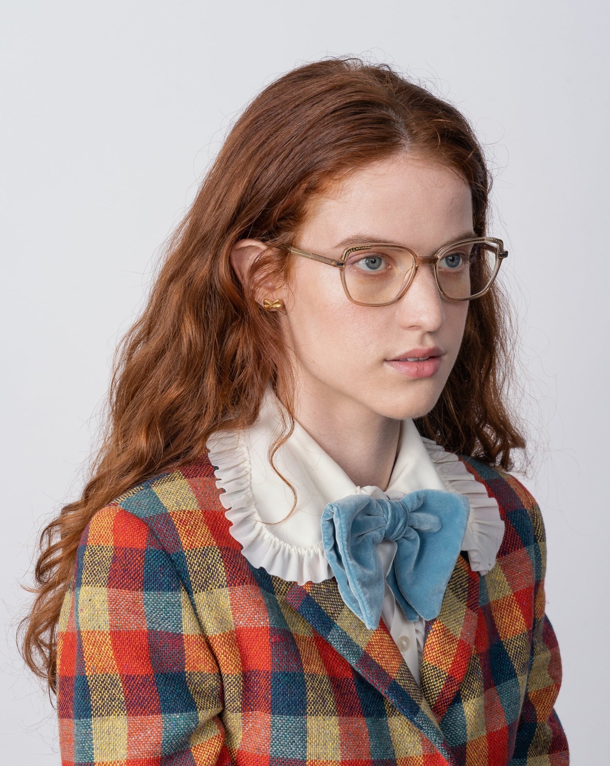A person with long, wavy auburn hair, wearing Mimosa by For Art's Sake® glasses with blue light filters, a white blouse with a ruffled collar, and a blue velvet bow tie is dressed in a multicolored plaid blazer. They are looking off to the side against a plain background.