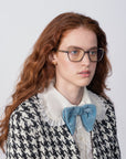 A woman with long, wavy auburn hair and Mimosa glasses by For Art's Sake® with prescription lenses is gazing slightly to the right. She is wearing a black and white houndstooth jacket over a white blouse with a ruffled collar, accessorized with a large light blue bow. The background is plain and light-colored.