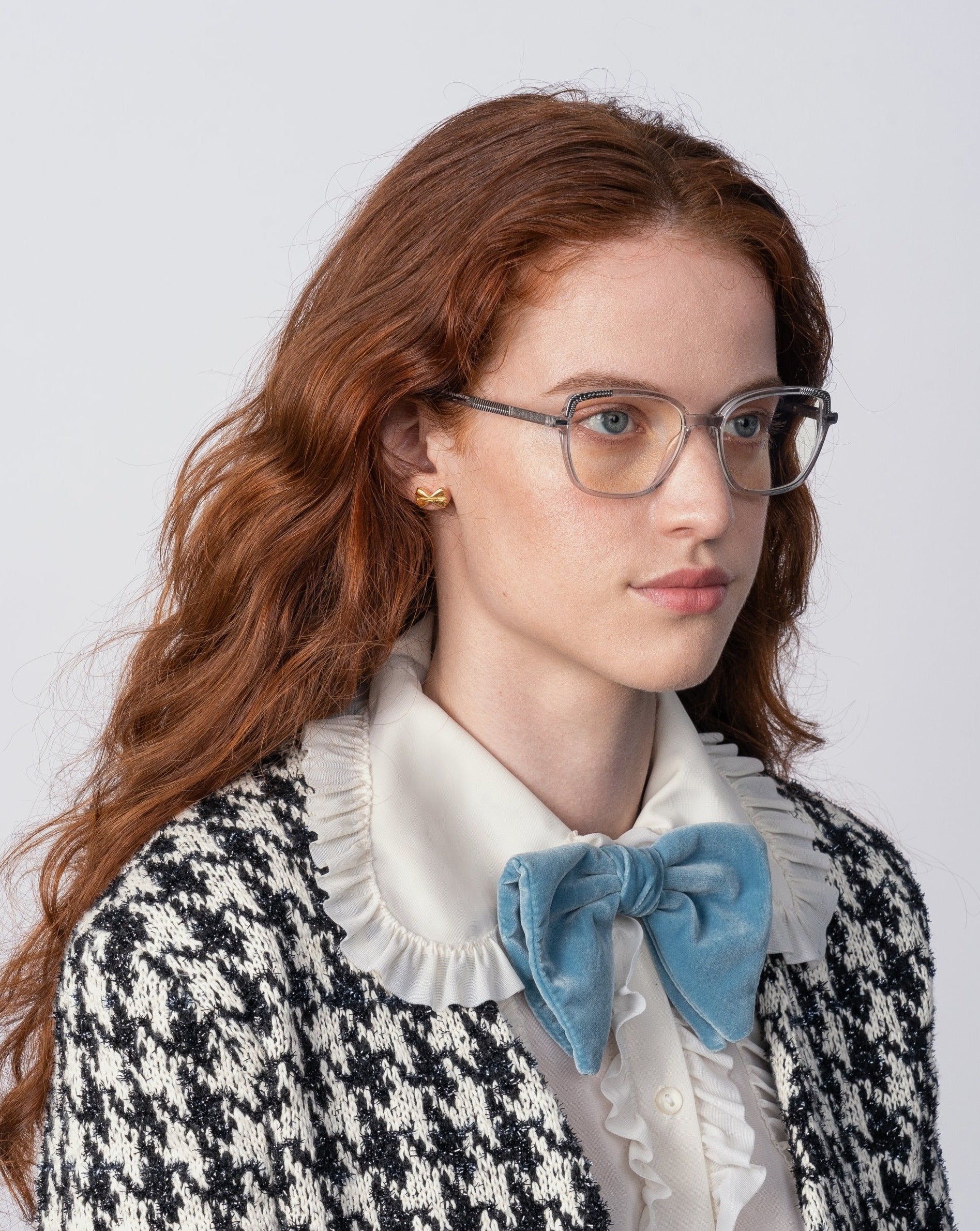 A person with long, red hair is wearing For Art&#39;s Sake® Mimosa prescription lenses and 18-karat gold-plated earrings. They are dressed in a black and white houndstooth patterned jacket, a white ruffled collar shirt, and a large blue bow at the neck. The background is plain and light-colored.
