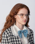 A person with long, red hair is wearing For Art's Sake® Mimosa prescription lenses and 18-karat gold-plated earrings. They are dressed in a black and white houndstooth patterned jacket, a white ruffled collar shirt, and a large blue bow at the neck. The background is plain and light-colored.