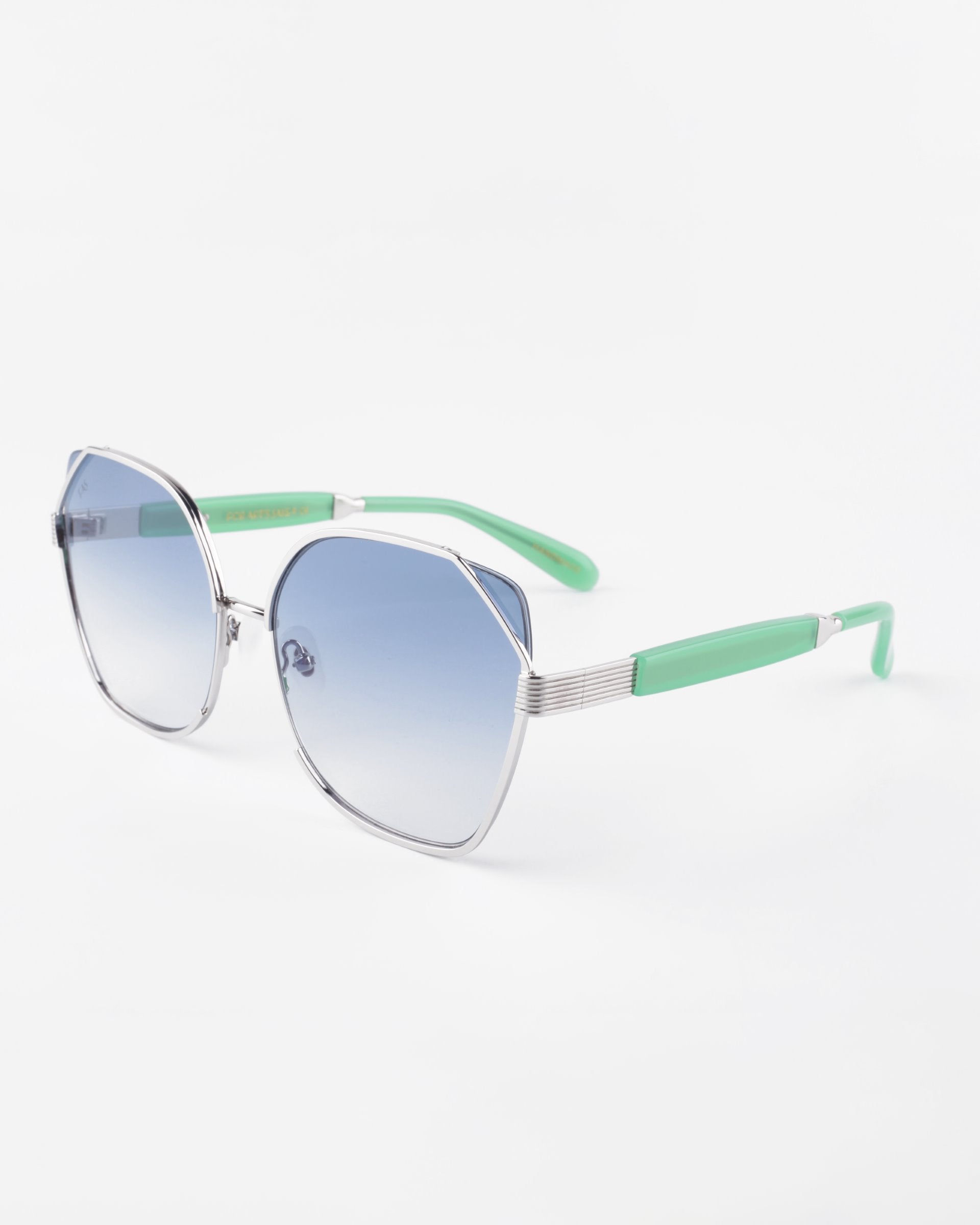 A pair of stylish For Art&#39;s Sake® Montage sunglasses featuring hexagonal frames with silver rims and light blue gradient lenses. The temples are mint green with silver accents near the hinges, crafted from gold-plated stainless steel. Ultra-lightweight nylon lenses provide 100% UVA &amp; UVB protection, creating a contemporary and fashionable look against a white background.