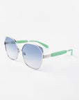 A pair of stylish For Art's Sake® Montage sunglasses featuring hexagonal frames with silver rims and light blue gradient lenses. The temples are mint green with silver accents near the hinges, crafted from gold-plated stainless steel. Ultra-lightweight nylon lenses provide 100% UVA & UVB protection, creating a contemporary and fashionable look against a white background.