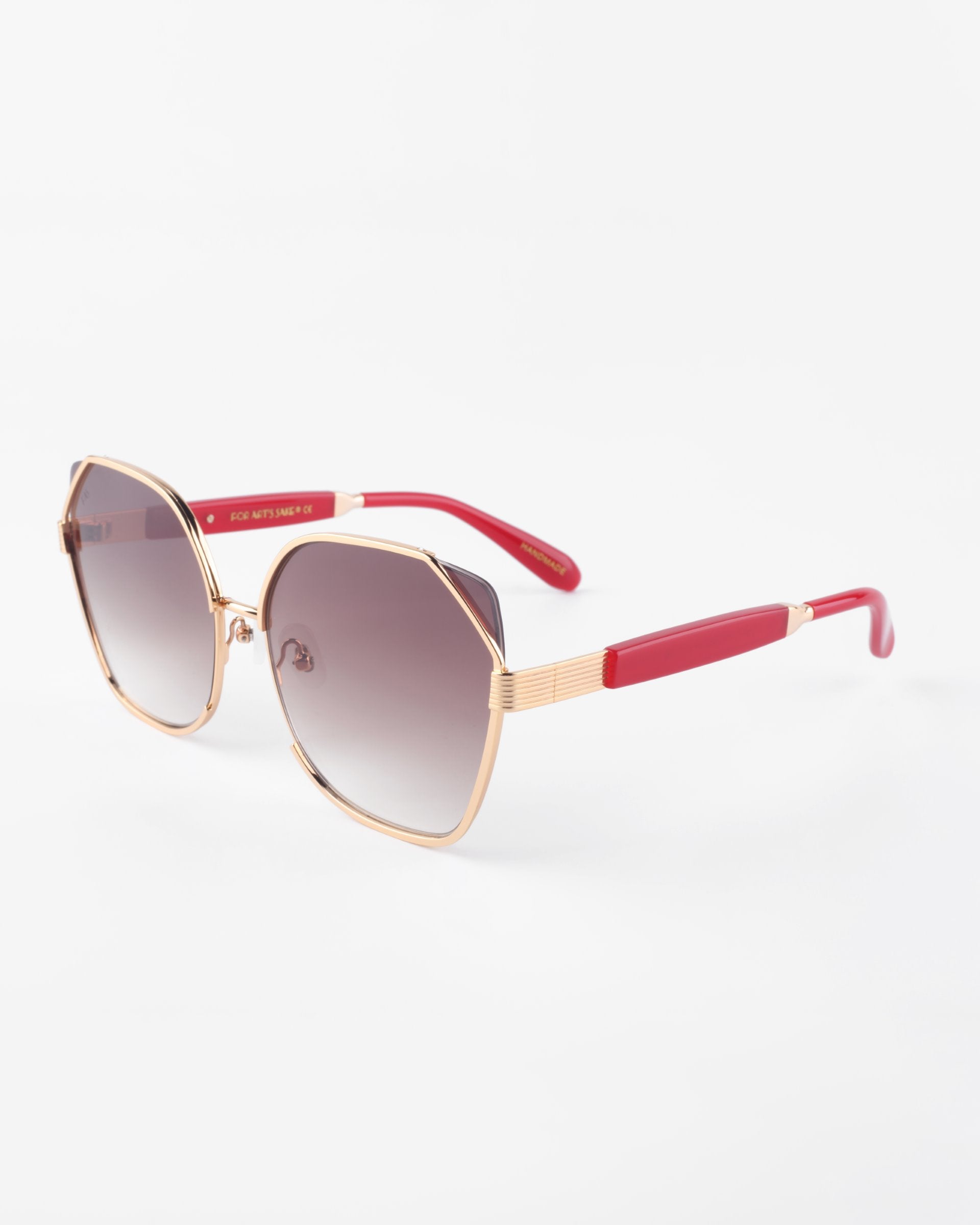 A pair of stylish Montage sunglasses by For Art&#39;s Sake® with a gold-plated stainless steel frame and hexagonal lenses, featuring red arms with gold accents. The ultra-lightweight nylon lenses are tinted in a gradient from dark to light, providing a sleek and modern look while offering 100% UVA &amp; UVB protection. The background is plain white.