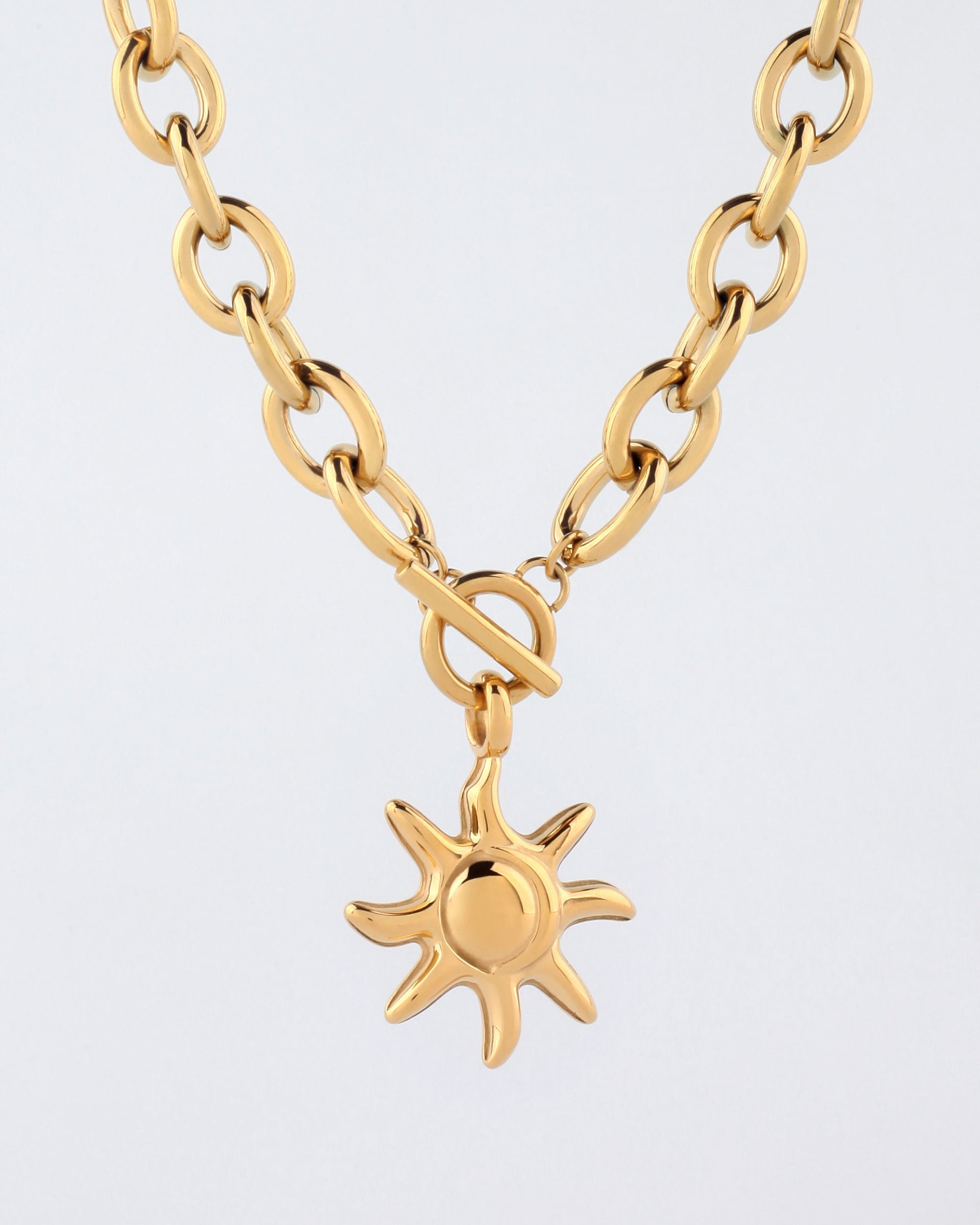 A close-up image of the For Art's Sake® Crescent Moon Necklace, featuring a celestial-inspired sun-shaped pendant with eight rays extending outward. The gold pendant is attached to a chunky chain and secured with a toggle clasp, all set against a plain white background.