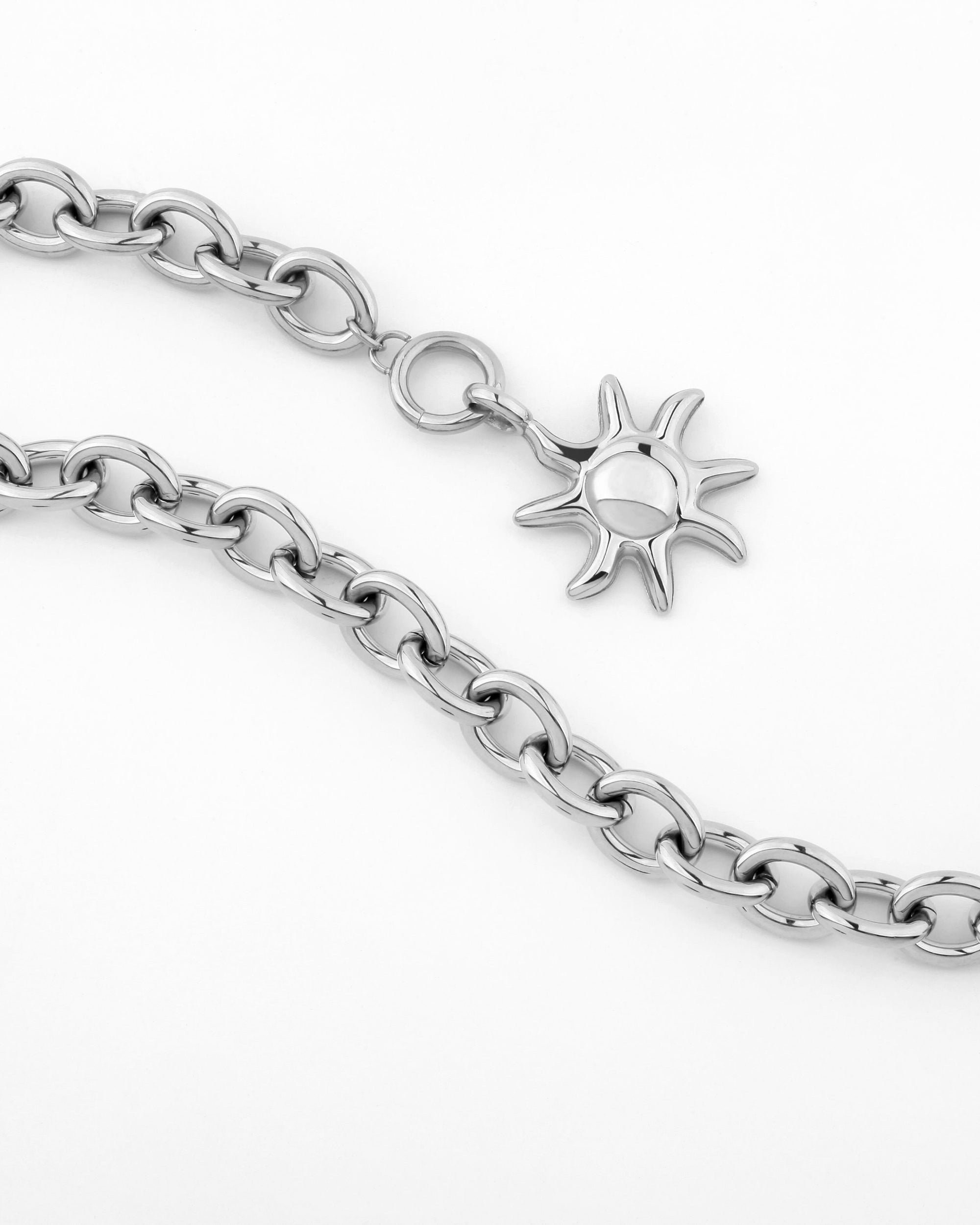 A silver chain necklace from For Art's Sake® with a celestial-inspired crescent moon-shaped charm attached. The charm features a central disc surrounded by pointed rays. The delicate chain has large, interlocking links, and the design boasts a polished, metallic finish on a plain background.