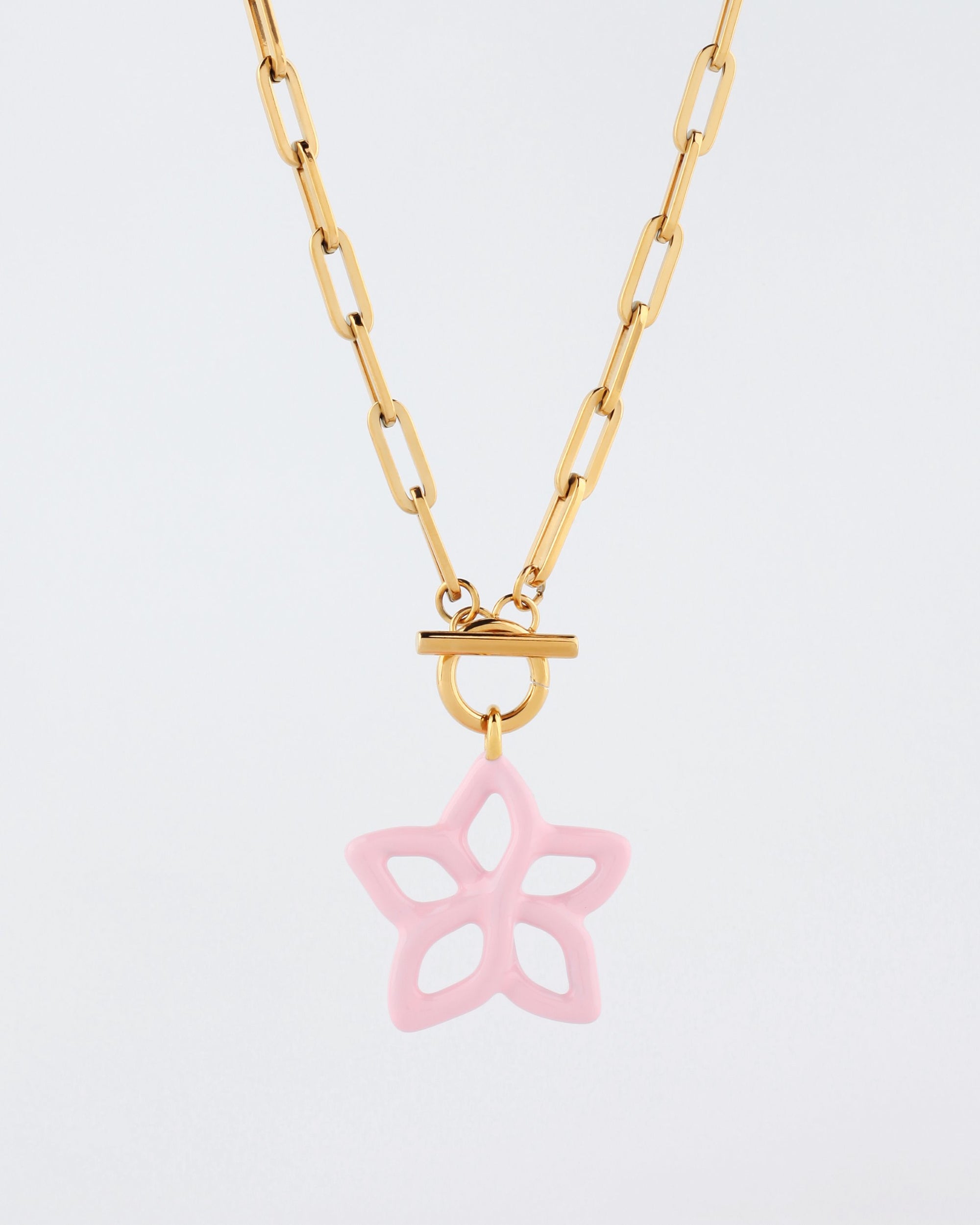 The Plumeria Necklace by For Art&#39;s Sake® features a gold chain with elongated links and a round toggle clasp that elegantly holds a pink star-shaped pendant. The five-pointed pendant boasts unique cut-out designs, giving it an airy and delicate look. Measuring 49cm in length, this piece is perfect for any occasion, especially against a plain white background.