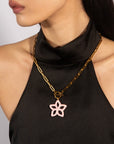 A close-up of a person wearing a sleeveless black top and the Plumeria Necklace from For Art’s Sake®, featuring a gold chain with a pink flower-shaped pendant. The 49cm-long necklace is the focal point, with the person's face partially visible, drawing attention primarily to the elegant accessory and top.