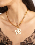 A close-up of a person wearing the Plumeria Necklace by For Art's Sake®, featuring a large, star-shaped pendant on a gold chain measuring 49cm in length. The individual is dressed in a black lace top, with long dark hair cascading down and their face partially visible.