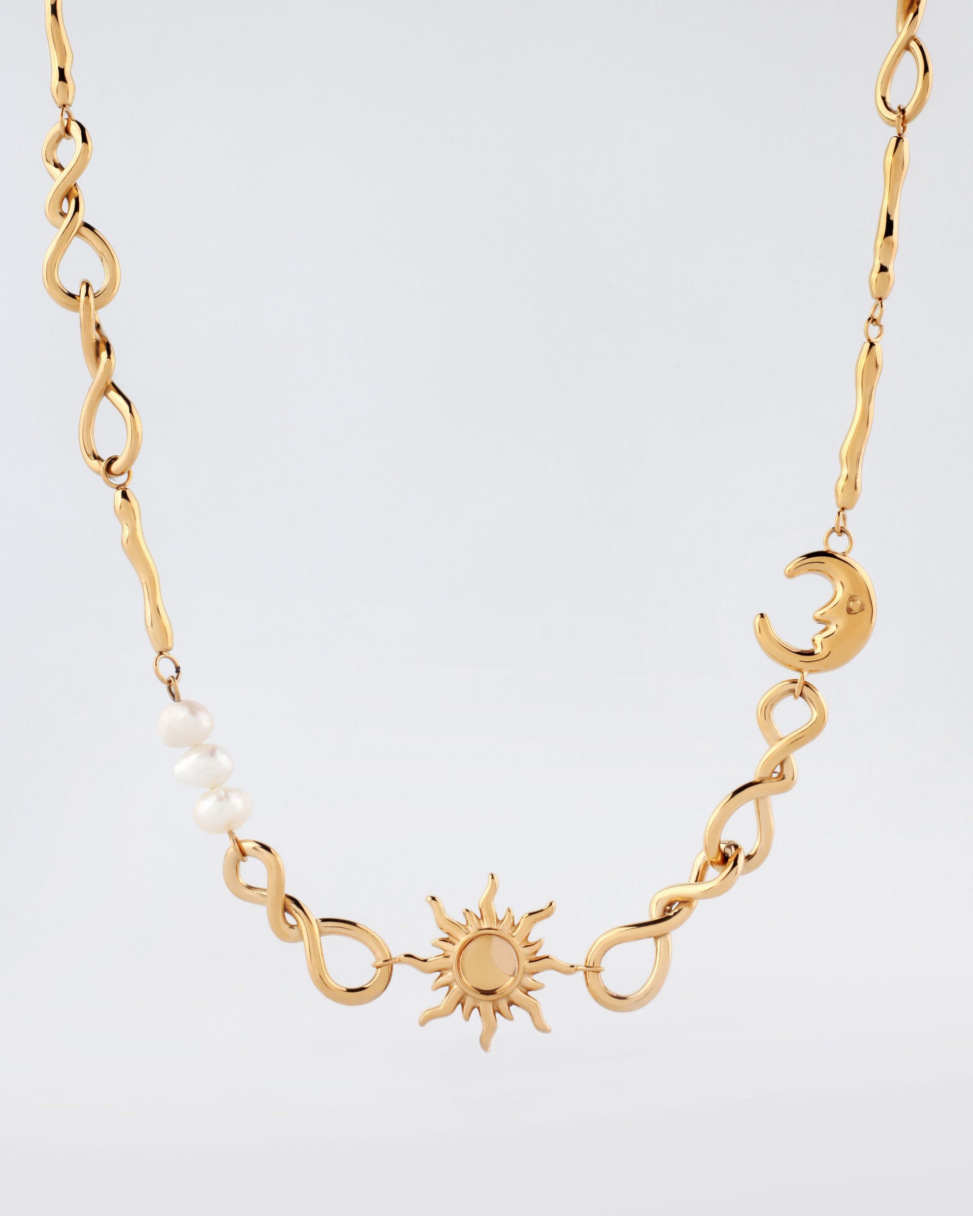 The Sloane Necklace by For Art's Sake® is a stunning gold piece, featuring a central sun pendant and a moon pendant on the right. The necklace showcases intertwined wavy chain links, adorned with several evenly spaced white pearls on the left side. This adjustable necklace is perfect for various measurements, all beautifully presented against a plain light-colored background.