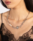 A person wears a black lace top and the distinct Sloane Necklace by For Art's Sake®. The necklace, adorned with silver-toned loops, a sun-shaped pendant, and several round white beads, drapes elegantly around their neck. An extension chain allows for an adjustable necklace length. The image focuses on the neck and chest area.