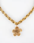 The Blossom Necklace by For Art's Sake® is a 46cm gold chain adorned with a detailed, five-petal flower pendant. The chain's interlocking links gracefully hold the pendant in the center, against a plain white background.