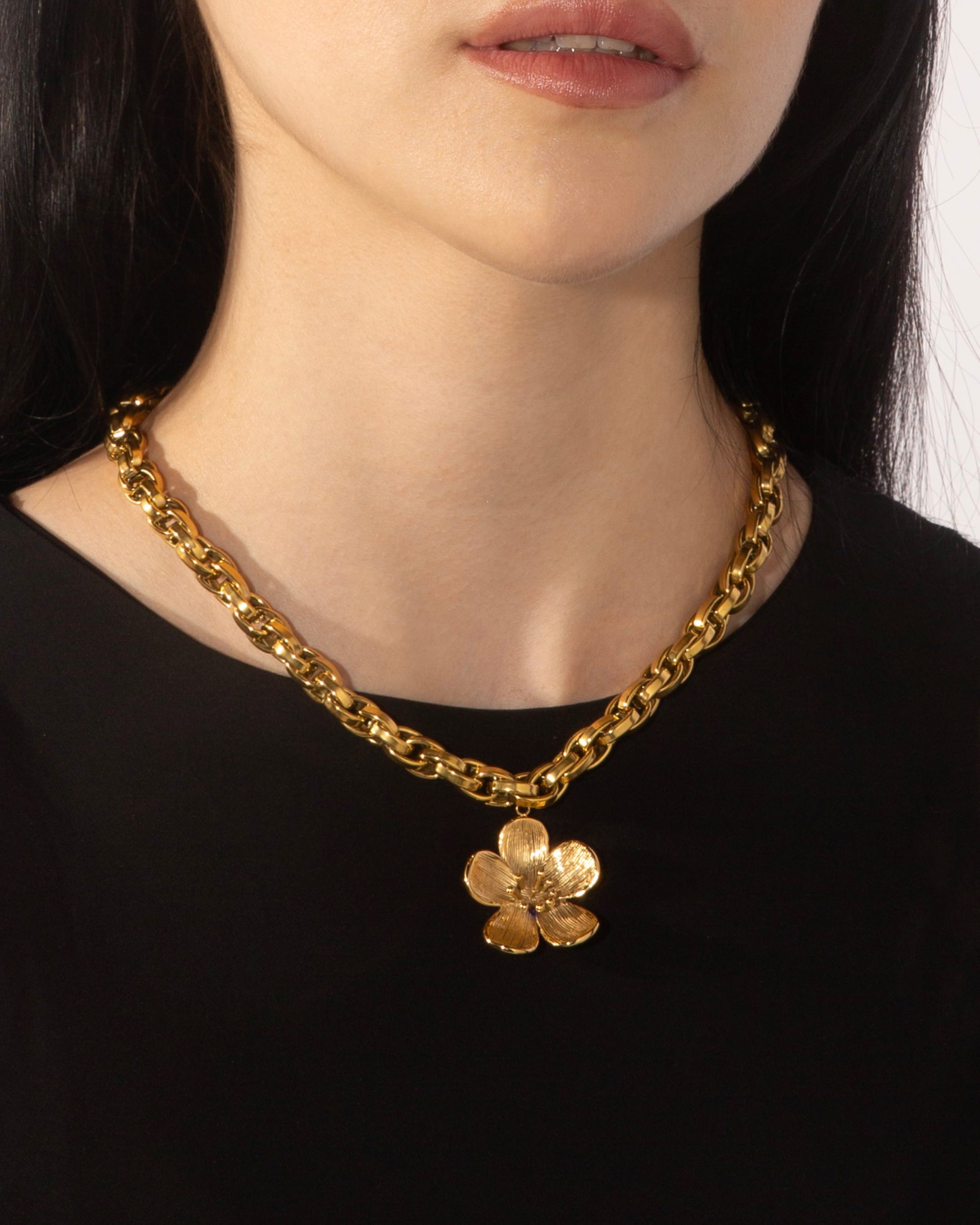 A person wearing a black top and the Blossom Necklace by For Art&#39;s Sake®, which is a chunky 46cm gold chain adorned with a large gold flower pendant. The image shows the person&#39;s neck and lower face.