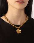 A person wearing a black top and the Blossom Necklace by For Art's Sake®, which is a chunky 46cm gold chain adorned with a large gold flower pendant. The image shows the person's neck and lower face.