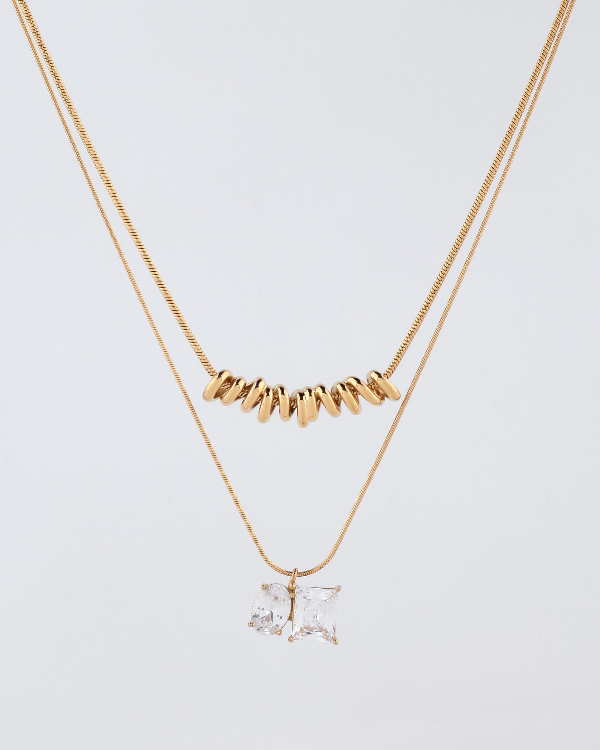 A minimalistic image features the Nora Necklace by For Art's Sake® against a plain white background. The top necklace, made up of small tubular gold beads, has an inner length that sits snugly around the neck. The bottom necklace is adorned with two square-cut crystal pendants—one clear and one with a hint of champagne color—and includes a 5cm extension chain for versatile styling.