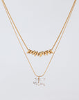 A minimalistic image features the Nora Necklace by For Art's Sake® against a plain white background. The top necklace, made up of small tubular gold beads, has an inner length that sits snugly around the neck. The bottom necklace is adorned with two square-cut crystal pendants—one clear and one with a hint of champagne color—and includes a 5cm extension chain for versatile styling.