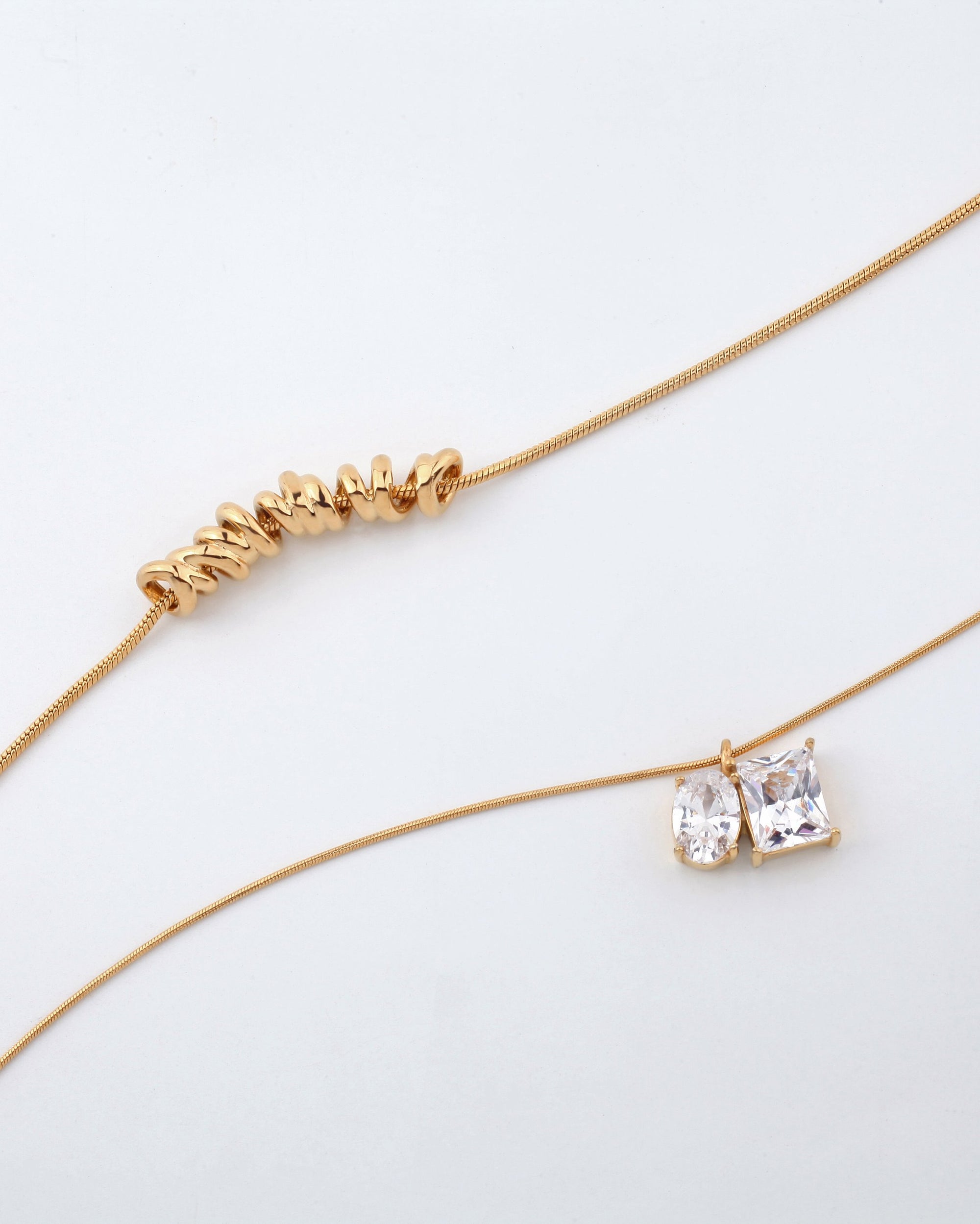 Displayed on a light background are two delicate gold necklaces from For Art&#39;s Sake®. The Nora Necklace, an adjustable piece with an extension chain for versatility, features an irregular row of small gold beads. The other necklace showcases two sparkling gemstones—a round and a square cut—hanging side by side as pendants.