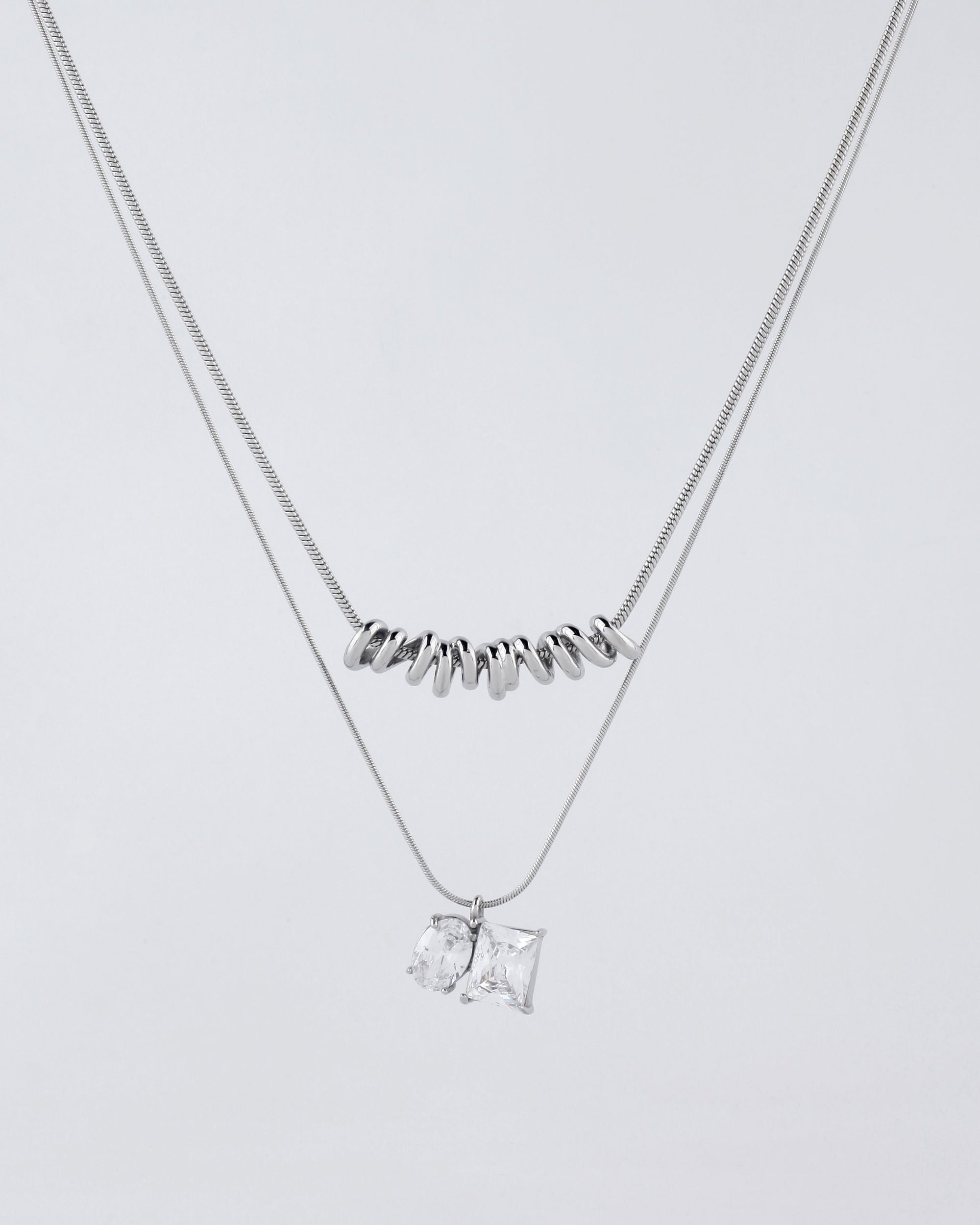 A photograph of two delicate Nora Necklaces by For Art&#39;s Sake®. The shorter Nora Necklace, adorned with a row of small cylindrical silver beads, and the longer one featuring two clear, square-cut pendants, create an elegant display against a plain light grey background. Their varied necklace lengths add to their charm and versatility.