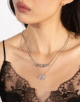 A woman wearing a black lace top and the Nora Necklace by For Art's Sake®, which includes two silver necklaces with pendants that feature extension chains for adjustable lengths. One necklace has a row of small, round beads, while the other showcases a detailed rectangular pendant. Her dark hair falls to her shoulders, adorned with a small earring.