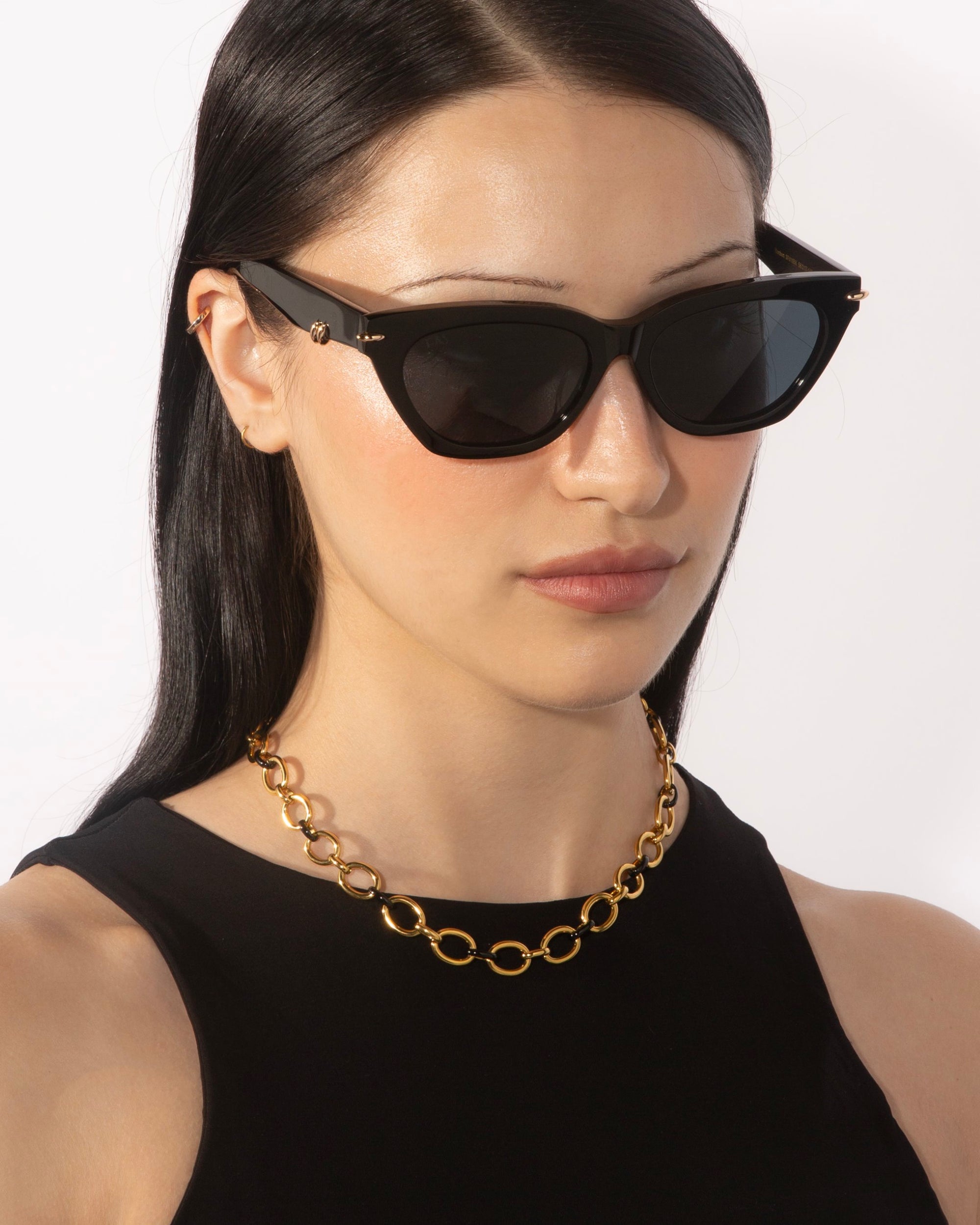 A person with long, dark hair wears cat-eye sunglasses and a black top, complemented by the bold Raya Necklace from the For Art&#39;s Sake® Love Letters collection. The background is a plain white wall.