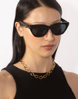 A person with long, dark hair wears cat-eye sunglasses and a black top, complemented by the bold Raya Necklace from the For Art's Sake® Love Letters collection. The background is a plain white wall.