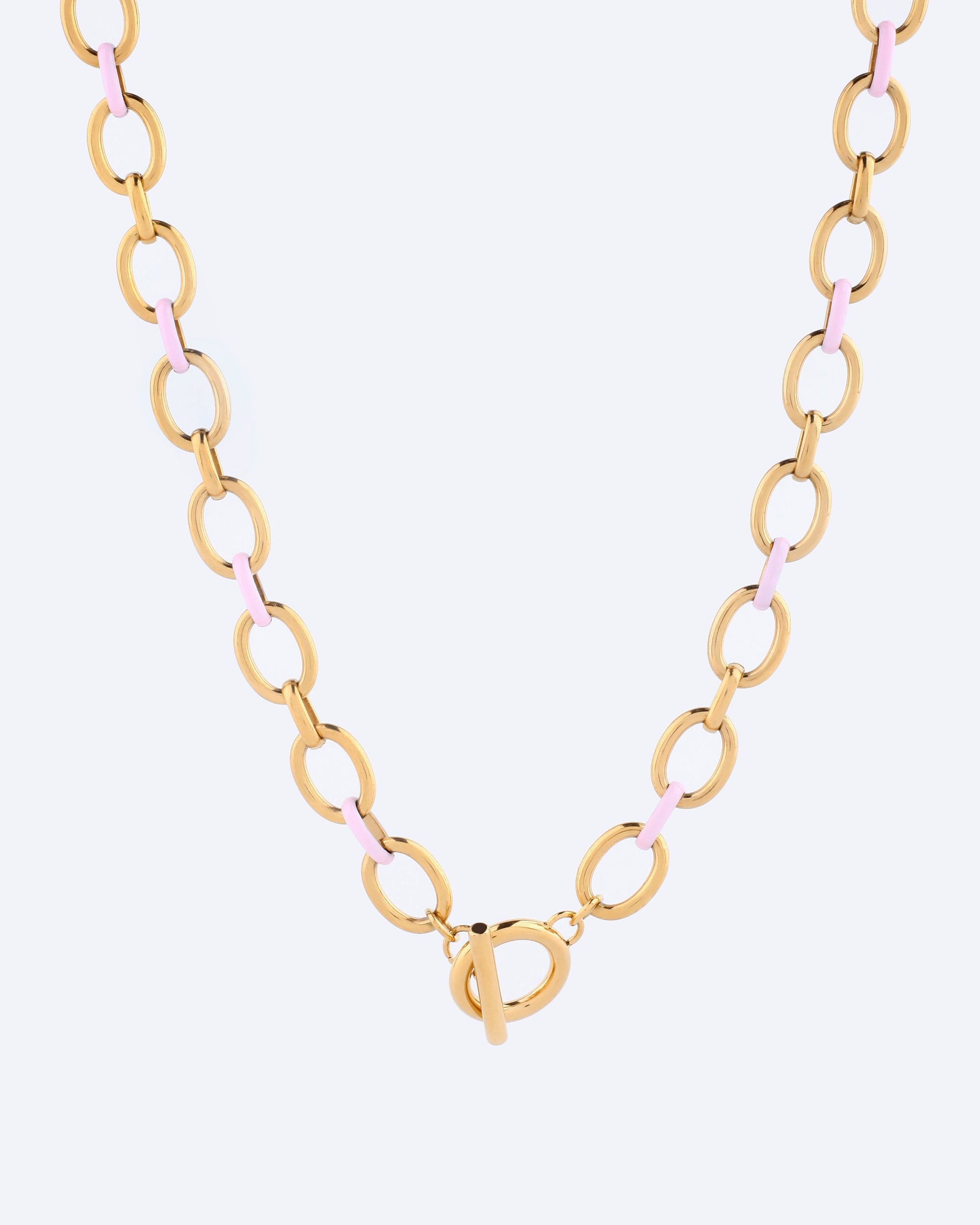 Discover the Raya Necklace by For Art&#39;s Sake®, a stunning gold creation from the Love Letters collection. It boasts a bold wide link chain with large oval links, highlighted by pink accents and finished with a central toggle clasp, beautifully set against a light backdrop.