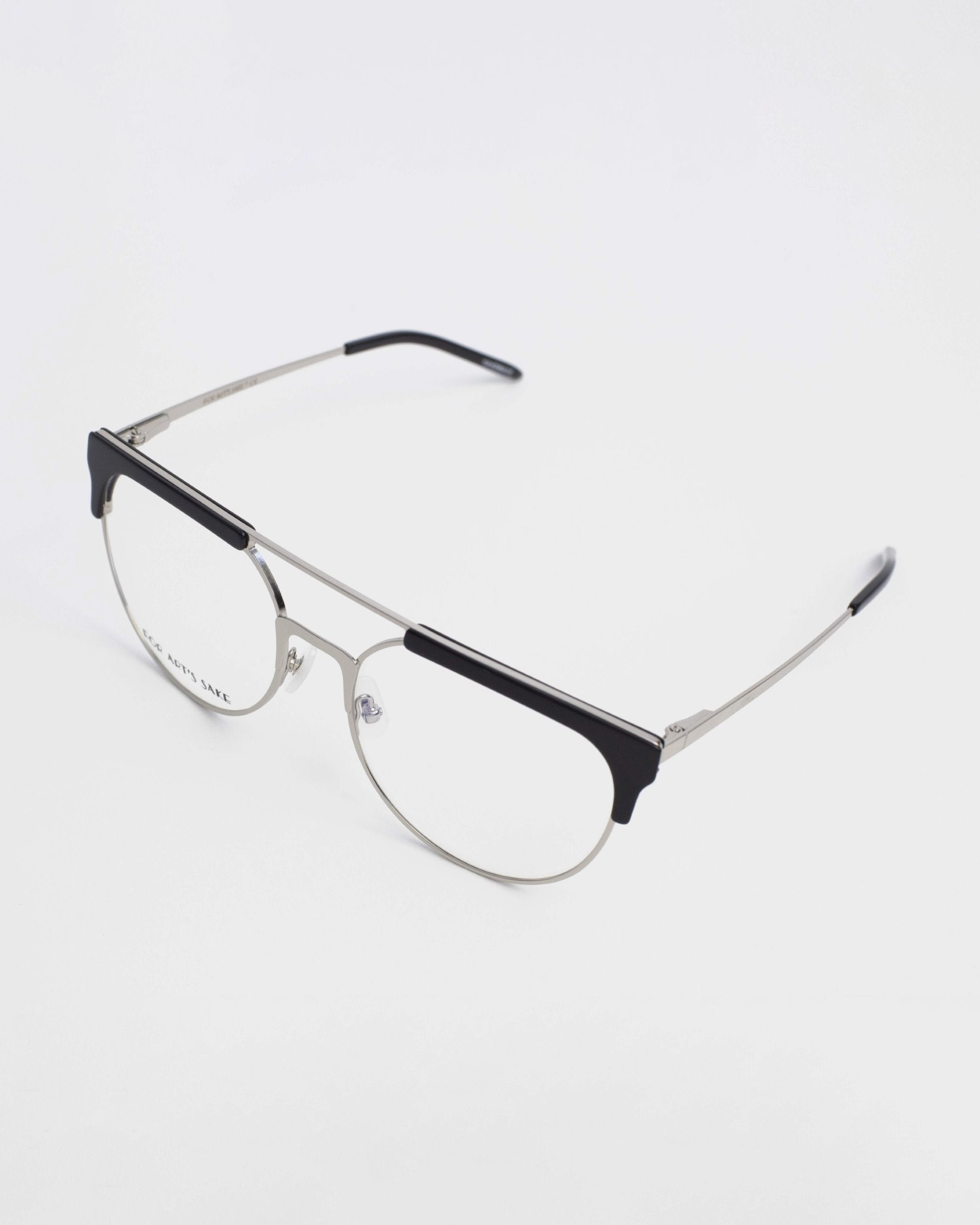 A pair of stylish Frosty prescription glasses by For Art&#39;s Sake® with a metallic frame and black accents on the top of the lenses, resting on a white surface. The design is minimalist with thin arms and clear lenses that include blue light filter technology.
