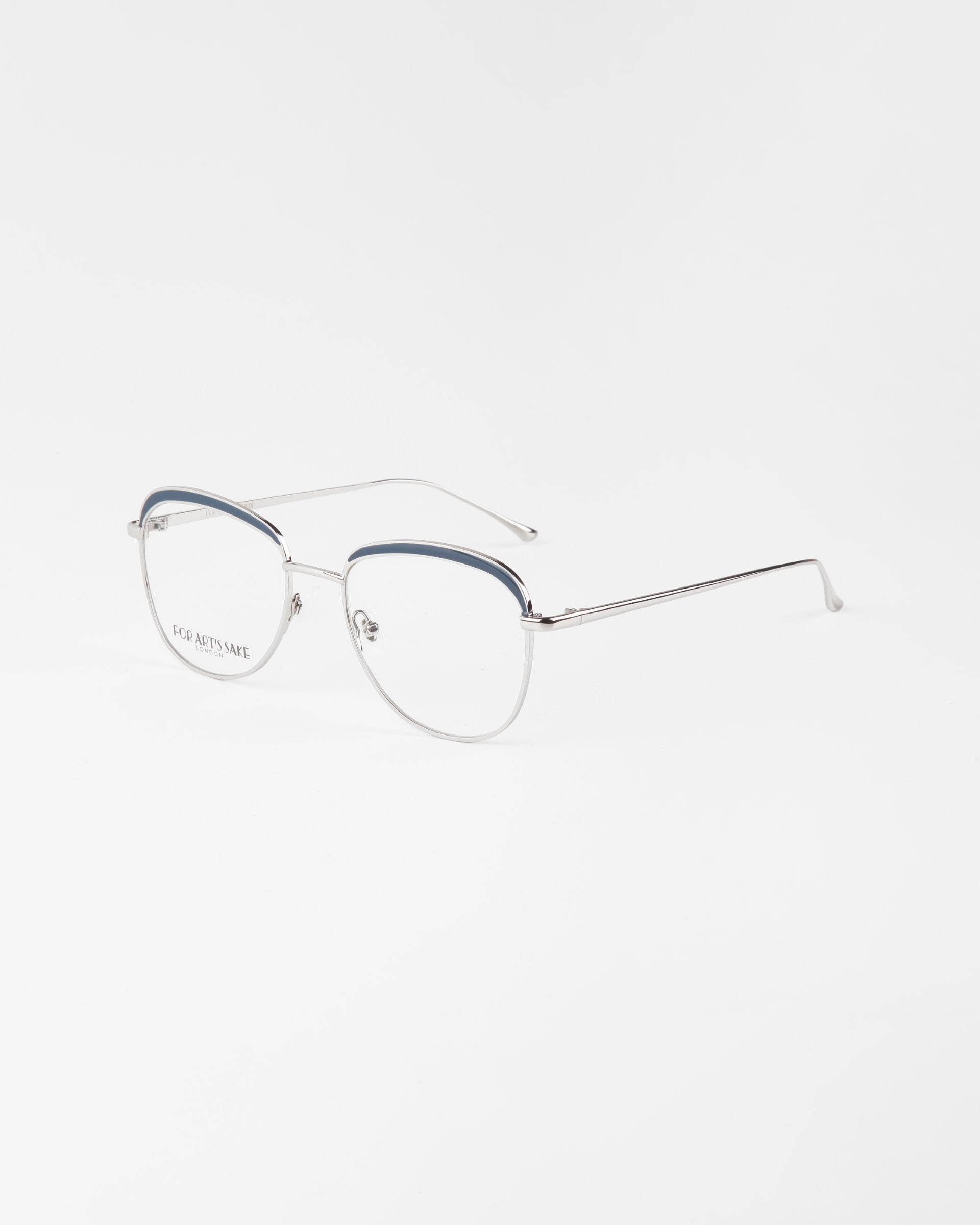 A pair of stylish, metal-rimmed eyeglasses called Smoothie from For Art&#39;s Sake® with a thin, silver frame and transparent lenses. The upper part of the rims is accented with a blue detail and includes a blue light filter. The glasses are positioned at an angle on a white surface.