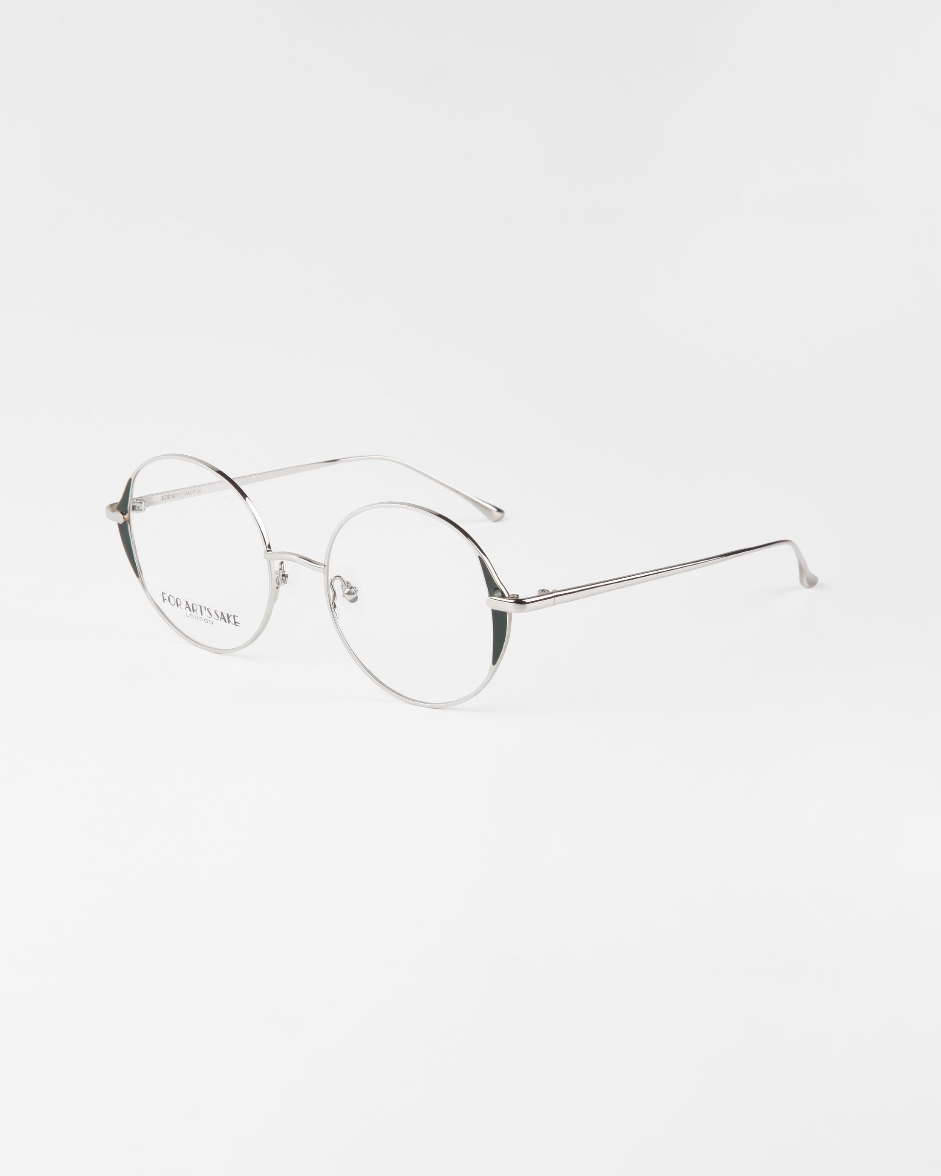 A pair of minimalist, round-framed eyeglasses with thin silver metallic frames and blue light filter lenses. The Kos glasses by For Art&#39;s Sake® rest on a white surface with a light grey background.