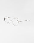 A pair of minimalist, round-framed eyeglasses with thin silver metallic frames and blue light filter lenses. The Kos glasses by For Art's Sake® rest on a white surface with a light grey background.