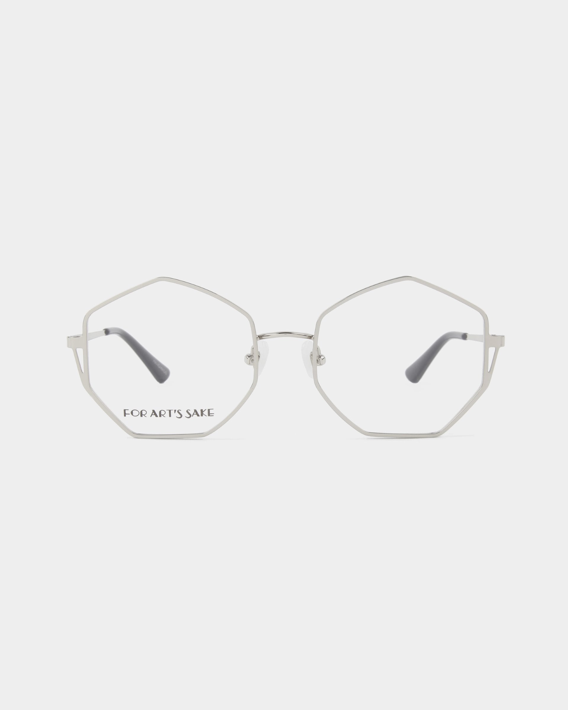 A pair of Antidote eyeglasses from For Art&#39;s Sake® with thin, 18-karat gold-plated octagonal frames and clear lenses. The bridge and temples are minimalistic, providing a modern and stylish look. The left lens displays the text &quot;FOR ART&#39;S SAKE&quot; in black. The background is plain white.