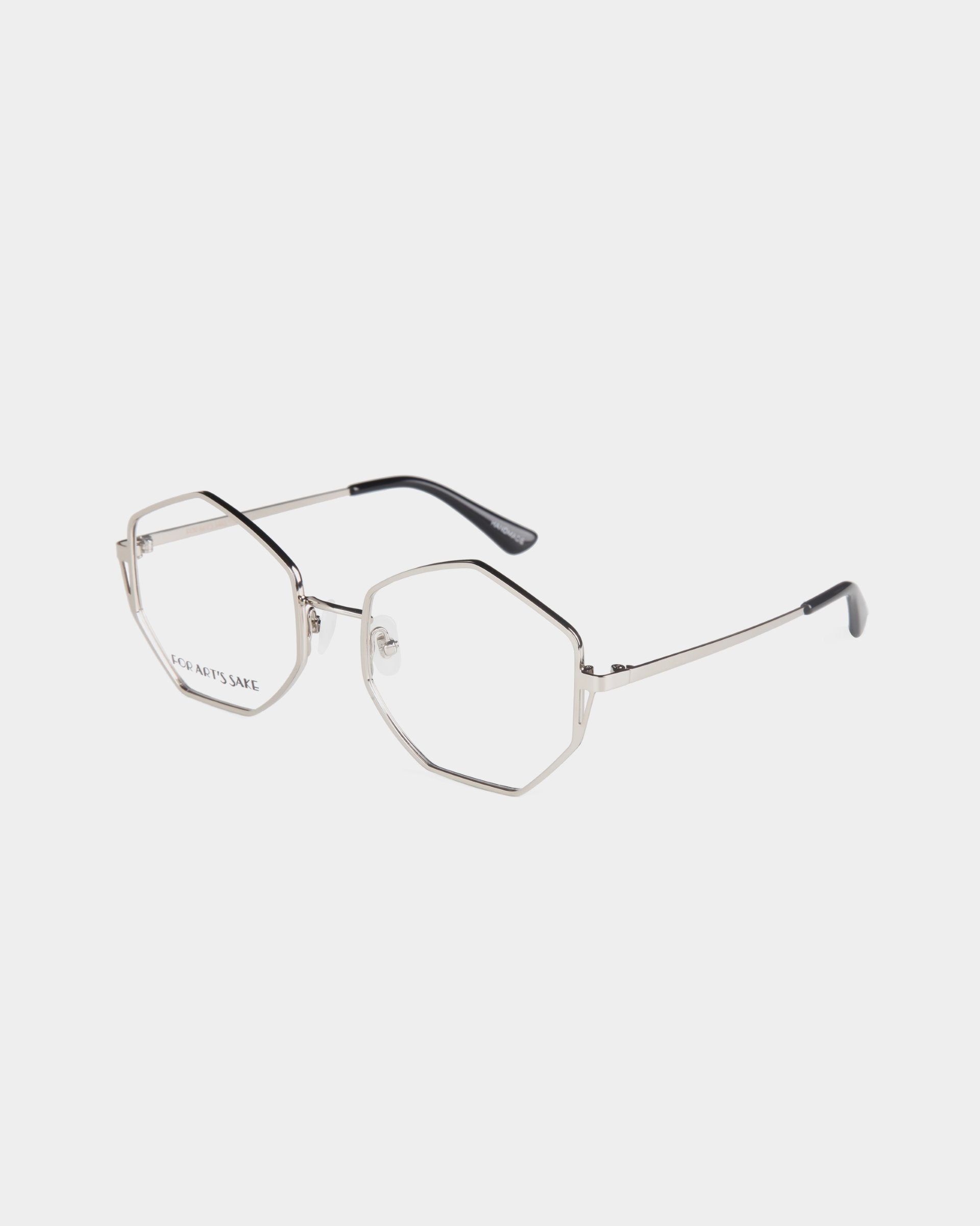 A pair of octagonal-shaped eyeglasses with thin silver metal frames and black tips on the arms. The lenses are clear and prescription-free, featuring a minimalist and modern design with adjustable jade nose pads. The words &quot;For Art&#39;s Sake® Antidote&quot; are visible on one of the lenses.