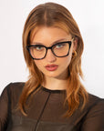 A person with long, reddish-brown hair is pictured against a plain background. They are wearing large, rectangular Dion eyeglasses by For Art's Sake® with tortoiseshell frames and gold hoop earrings. They have a neutral expression and are dressed in a sheer black top layered over a black garment.