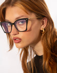 A person with long, reddish-brown hair and tortoiseshell frames is looking intently at the camera. They wear large black For Art's Sake® Dion glasses with a gold detail on the arms and gold hoop earrings. With a light skin tone and dressed in a black top, they stand against a plain white background.