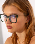 A person with long, light brown hair is wearing large, black-framed eyeglasses with gold details featuring a blue light filter from For Art's Sake®'s Dion collection. They also wear gold hoop earrings and a gold chain necklace. The person is gazing slightly to the side with a neutral expression against a white background.