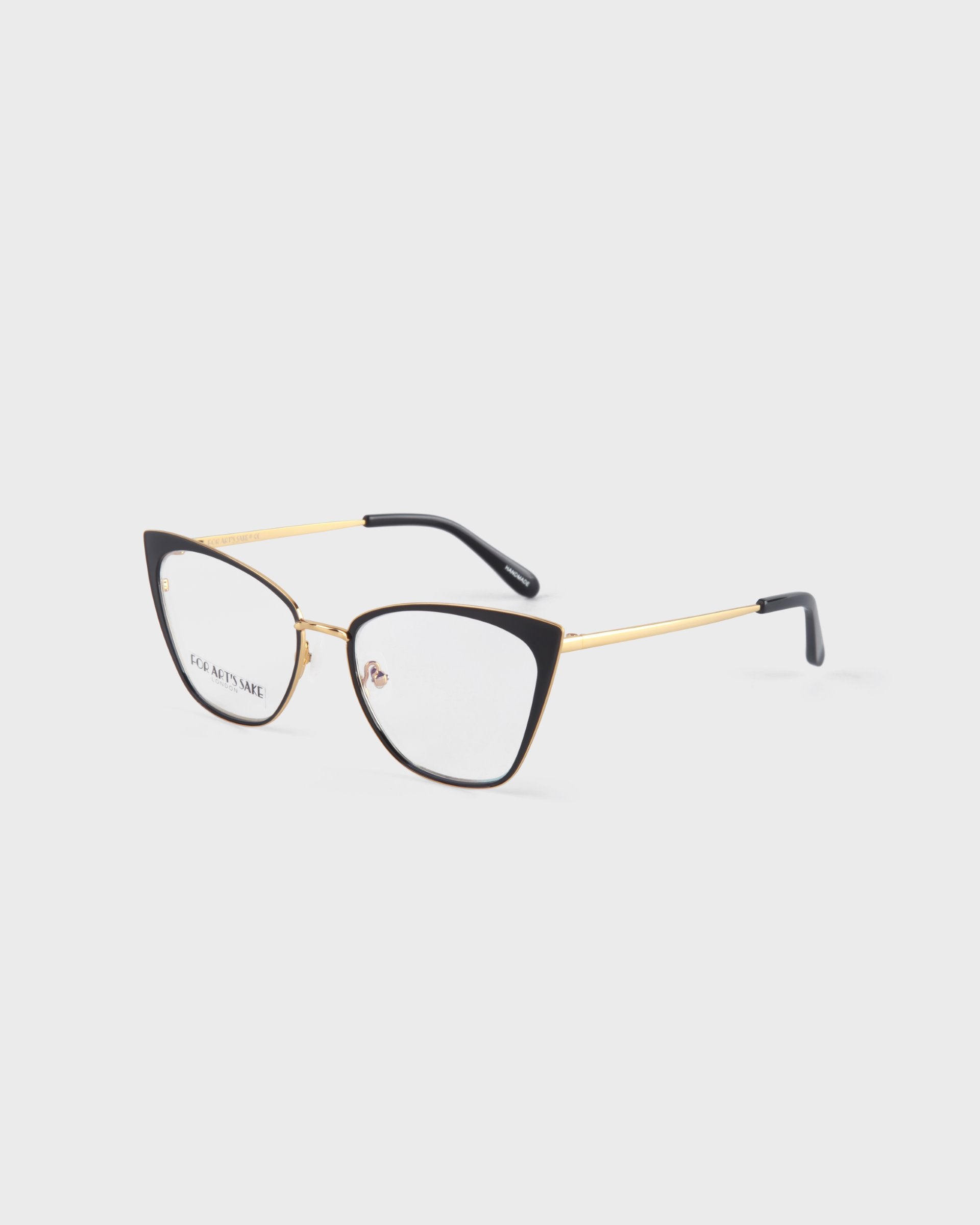 A pair of stylish Stella Two eyeglasses by For Art&#39;s Sake® with a black cat-eye frame and 14kt gold plated arms. The lenses are clear, and the tips of the arms are black. The brand name is visible on the inside of the left lens. Prescription services available. The background is white.