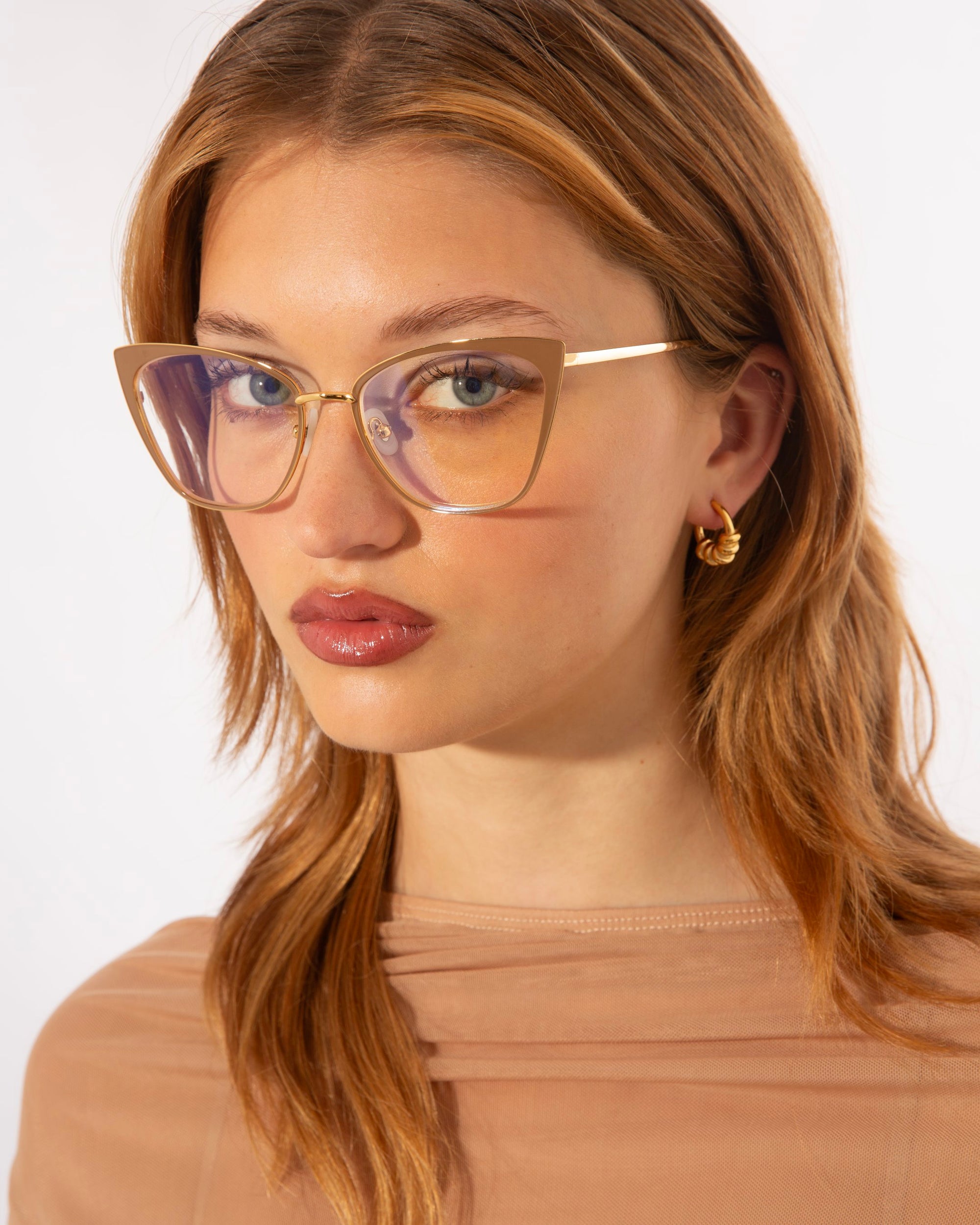 A young woman with light brown hair and wearing glasses looks directly at the camera. Her *Stella Two* glasses by *For Art's Sake®*, adorned with a golden frame and featuring blue light filter lenses, complement her small 14kt gold-plated hoop earrings. She is dressed in a sheer beige top, standing against a plain white background.