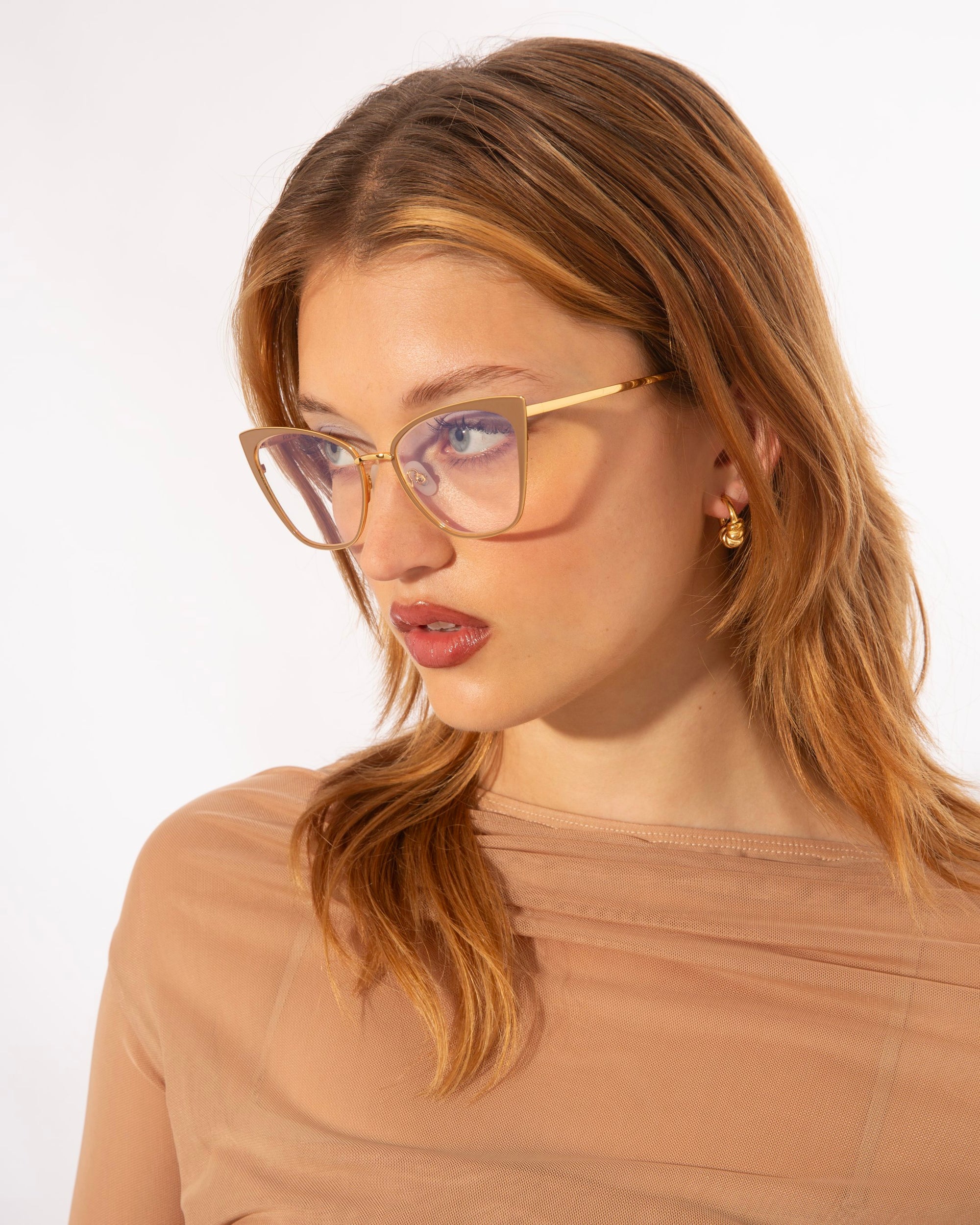 A woman with light brown hair wearing stylish, oversized glasses with a Blue Light Filter and small 14kt gold plated hoop earrings looks slightly to the side. She is dressed in a sheer, beige top. She is wearing Stella Two by For Art's Sake®. The background is plain white, giving a clean and minimalist aesthetic to the image.