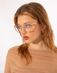 A woman with light brown hair wearing stylish, oversized glasses with a Blue Light Filter and small 14kt gold plated hoop earrings looks slightly to the side. She is dressed in a sheer, beige top. She is wearing Stella Two by For Art's Sake®. The background is plain white, giving a clean and minimalist aesthetic to the image.