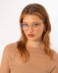 A person with long, light brown hair wears gold-rimmed For Art's Sake® Stella Two glasses featuring Blue Light Filter technology and a sheer, beige-colored top. They have a neutral expression on their face and are posed against a plain white background.