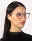 A person with long dark hair wears the chic Stella Two oversized eyeglasses by For Art's Sake®, featuring a cat-eye frame with gold accents and semi-transparent temples. They are dressed in a black top against a plain white background, looking slightly to the side.