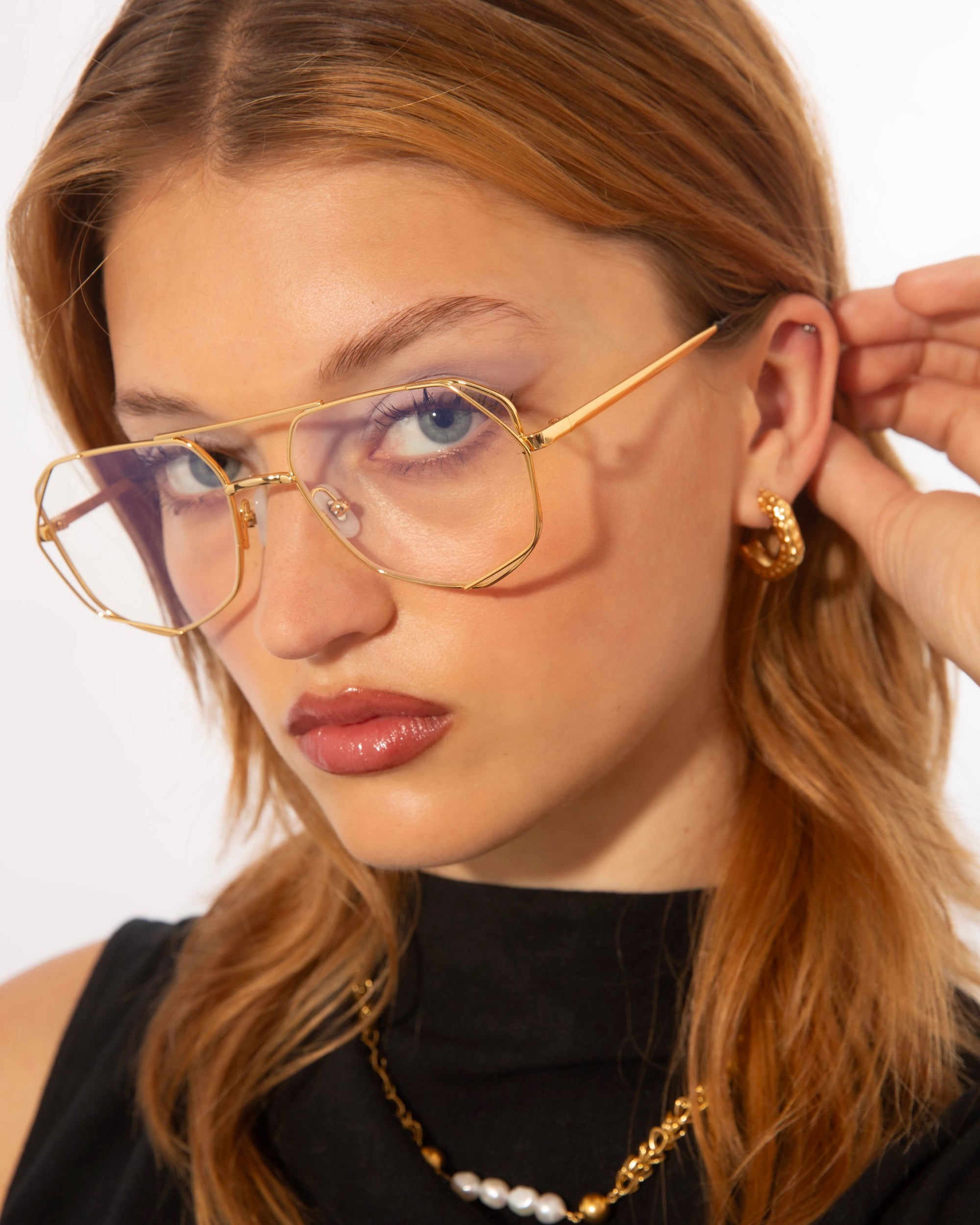 A woman with light brown hair wears oversized, gold-framed Genius Two glasses from For Art&#39;s Sake® with blue light filter lenses and gold hoop earrings. She is dressed in a black top with a necklace featuring pearls. Her hand is gently touching her ear, and she gazes directly at the camera. The background is plain white.