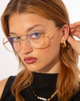 A woman with light brown hair wears oversized, gold-framed Genius Two glasses from For Art's Sake® with blue light filter lenses and gold hoop earrings. She is dressed in a black top with a necklace featuring pearls. Her hand is gently touching her ear, and she gazes directly at the camera. The background is plain white.