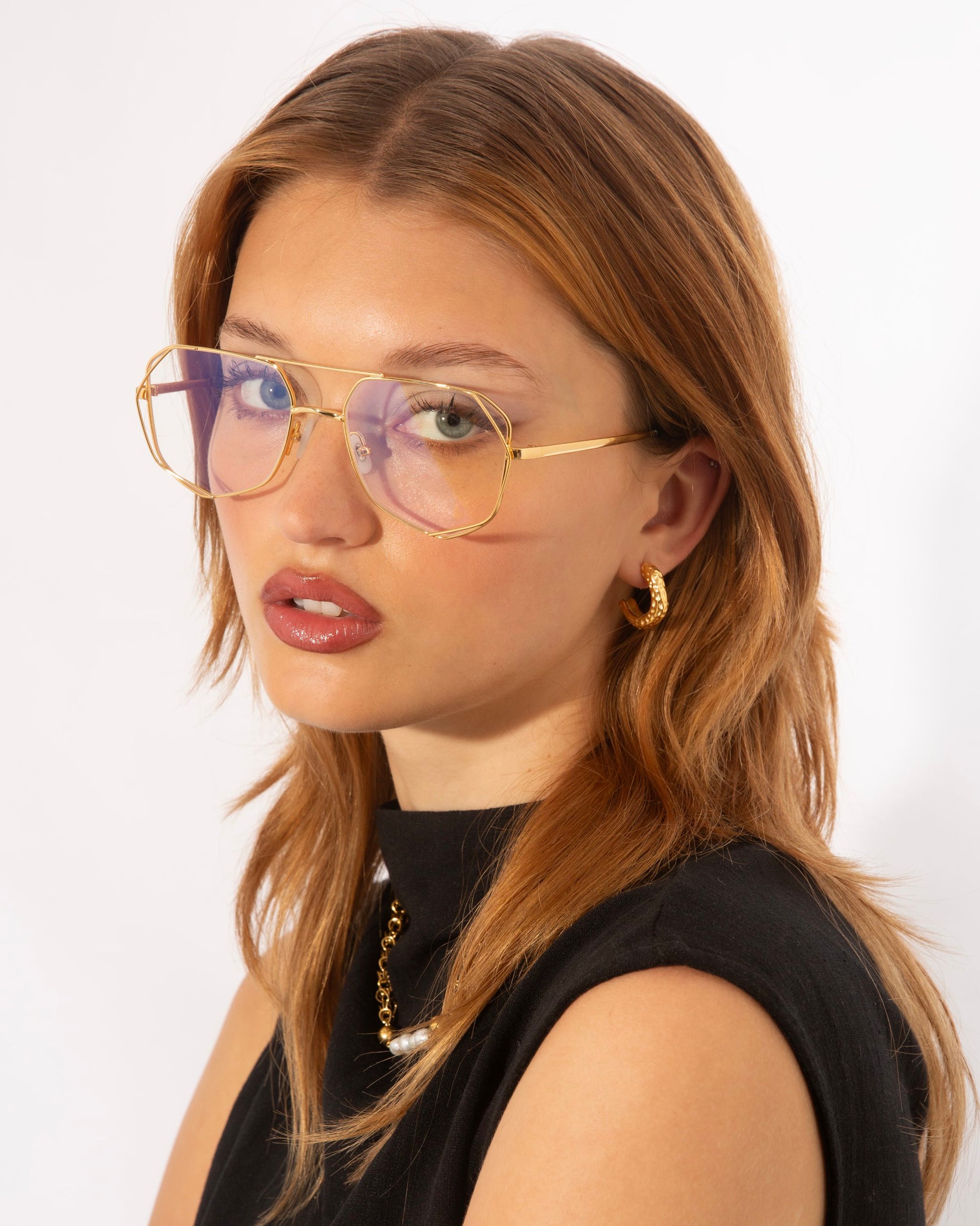 A person with shoulder-length, light brown hair wears large, gold-rimmed glasses featuring UV protection lenses with a purple tint. They have gold hoop earrings and a necklace and are dressed in a sleeveless black top. The person looks directly at the camera against a plain white background, wearing For Art's Sake® Genius Two glasses.