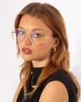 A person with shoulder-length, light brown hair wears large, gold-rimmed glasses featuring UV protection lenses with a purple tint. They have gold hoop earrings and a necklace and are dressed in a sleeveless black top. The person looks directly at the camera against a plain white background, wearing For Art's Sake® Genius Two glasses.