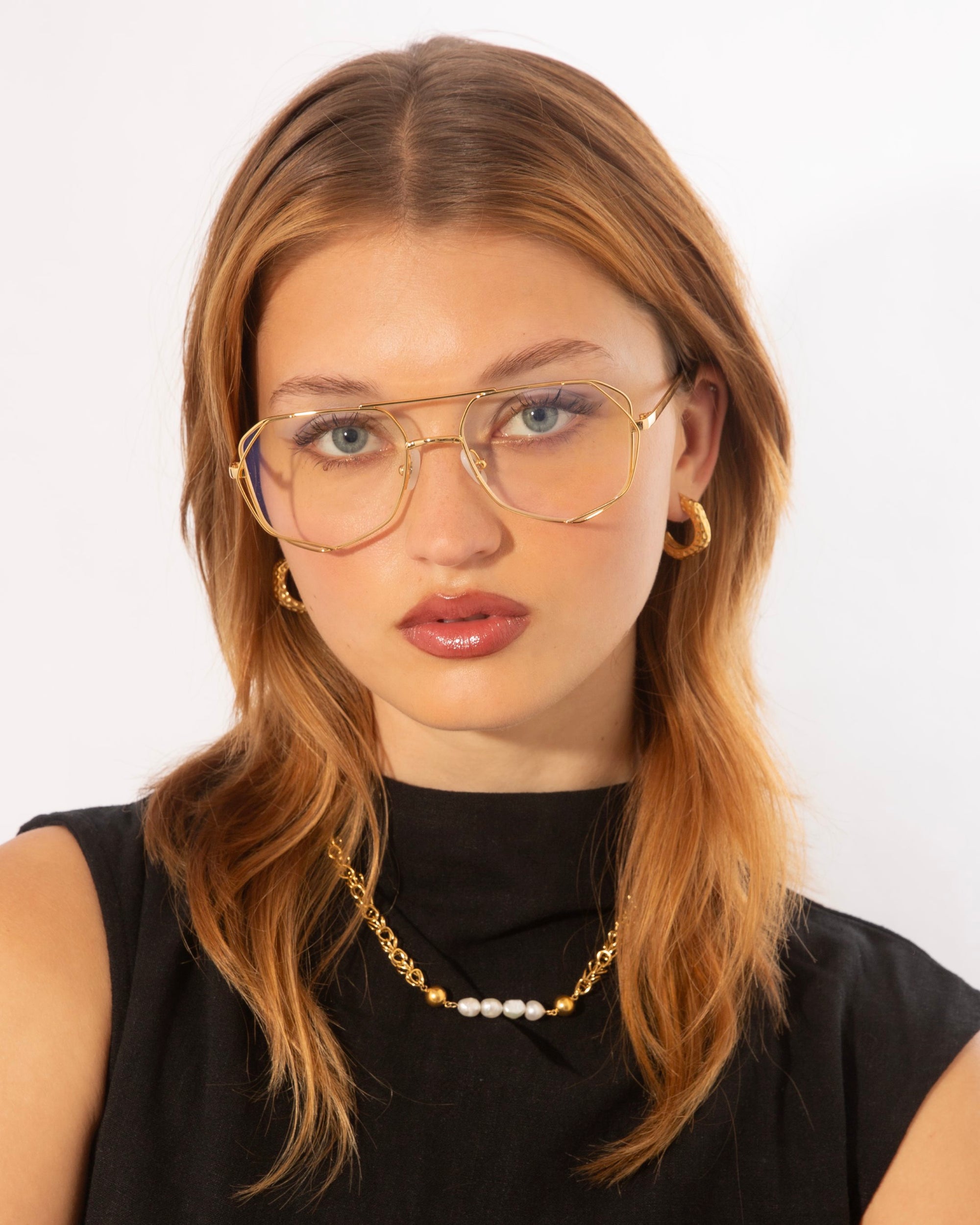 A young woman with straight reddish-brown hair and wearing For Art's Sake® Genius Two glasses, looks at the camera. She is dressed in a black sleeveless top and accessorized with gold hoop earrings and a gold necklace with white beads. The background is plain white.