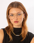 A young woman with straight reddish-brown hair and wearing For Art's Sake® Genius Two glasses, looks at the camera. She is dressed in a black sleeveless top and accessorized with gold hoop earrings and a gold necklace with white beads. The background is plain white.